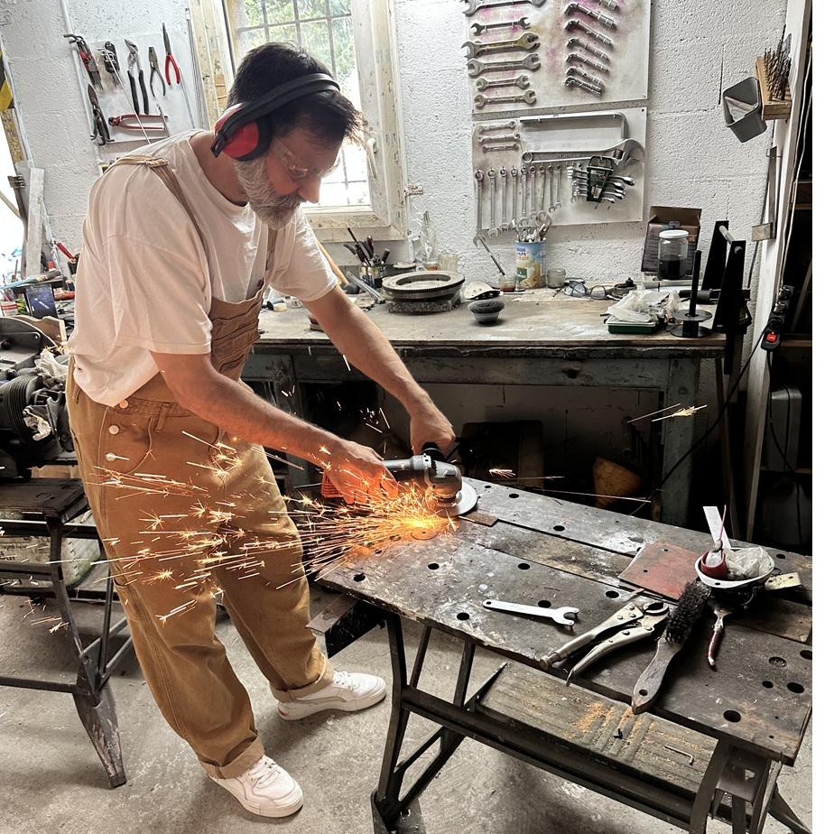 Laurent au travail dans son atelier, meulage d'une pièce métallique en gerbe d'étincelles, établi couvert d'outils